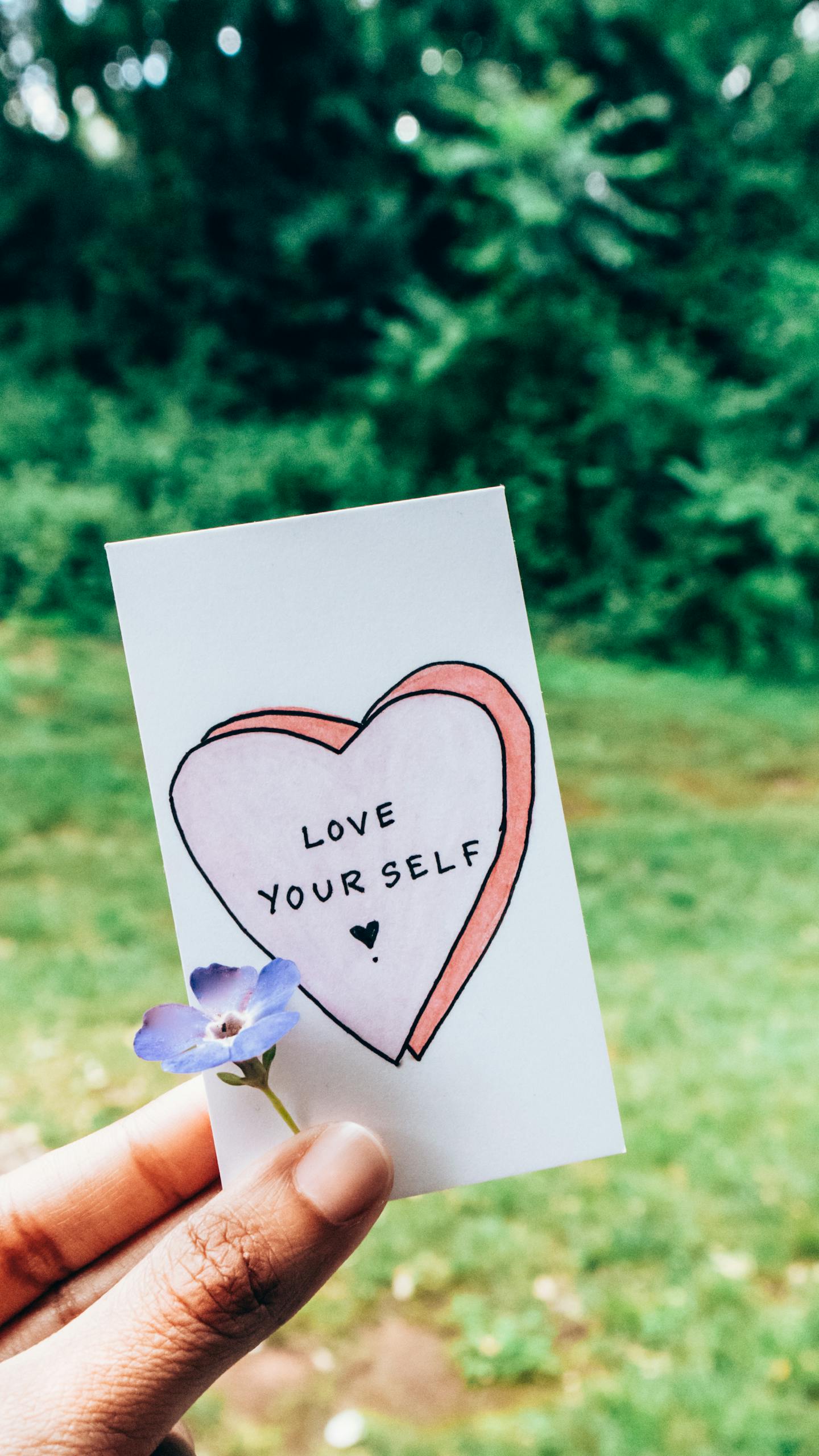 A hand holds a card with 'Love Yourself' next to a small flower outdoors.
