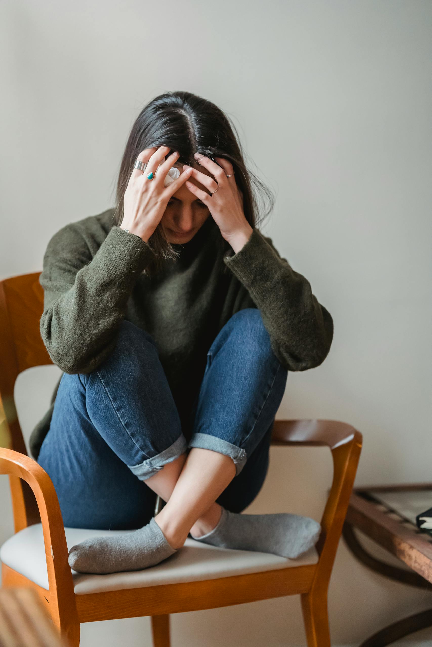 Full body of unhappy young ethnic lady with dark hair in casual clothes touching head and looking down sadly while sitting on chair with crossed legs