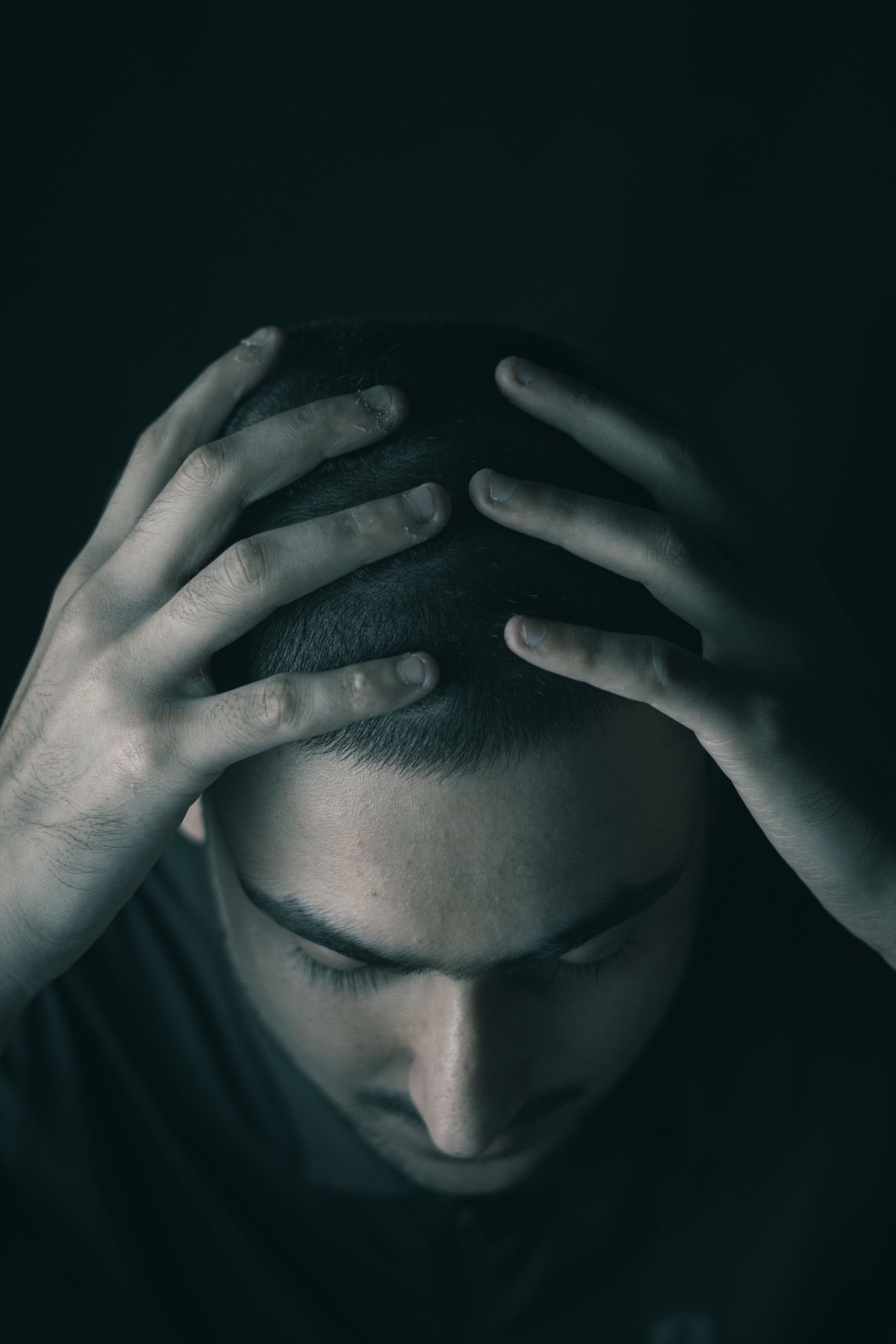 A man sitting in a dark room, hands on head, conveying stress and depression.